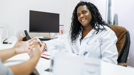 A dark-skinned trans woman sitting indoors at a desk, grinning happily as she looks at the individual sitting on the other side of the desk, who is blurred in the foreground. The woman is a doctor, wearing a white medical coat, with long, curly black hair and brown eyes. The person in the foreground, her patient, is a lighter-skinned woman, who is gesturing while speaking, wearing a long-sleeved grey shirt. The interior of a medical office, including a computer monitor and white walls, is visible in the blurred background.