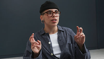 A non-binary person sitting indoors, gesturing as they speak