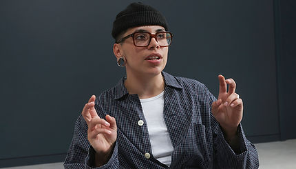 A non-binary person sitting indoors, gesturing as they speak