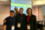 Four people posed smiling with a Powerpoint presentation on display in the background