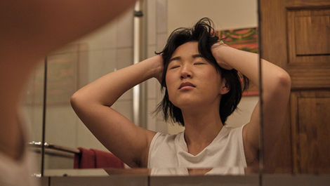An Asian non-binary trans man standing indoors in a dimly-lit bathroom. His eyes are closed and he is holding his head with both hands, as if massaging his temples or displaying stress. He is wearing a sleeveless white shirt. He has shoulder-length, wavy black hair and tan skin. A shower, wooden door, white wall, and tiled wall are visible in the blurred background.