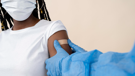 A Black girl sitting indoors, wearing a white face mask and a short-sleeved white shirt. The shirt sleeve which is visible has been rolled up, and a pair of blue gloved hands are pressing a band-aid to the girl's upper arm, implying she has just received a vaccine. She has long, braided black hair, and is wearing a pair of gold hoop earrings.