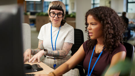 A gender non-conforming, light-skinned woman sitting at a desk, looking towards a computer and gesturing as she speaks. A darker-skinned woman is sitting directly in front of the computer, using it while listening to the other woman's instructions. The speaking woman has short, black hair partly bleached blonde, and is wearing a white shirt, patterned tan trousers, black-rimmed glasses, and a blue lanyard. The woman working on the computer has curly, dark brown hair and is wearing a maroon shirt and a blue lanyard. An office space is visible in the blurred background.