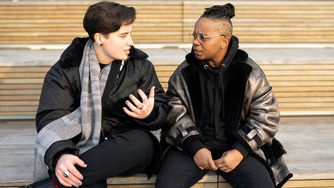 Two transmasculine people sitting outdoors on a wooden stairwell, looking at each other and speaking. The person on the left is light-skinned with short, black hair, wearing a padded black jacket, a grey scarf, black trousers, and rings. Their nails are painted black. The person on the right is a darker-skinned person with partly-shaved black hair, styled into dreadlocks and tied up into a bun. They are wearing a metallic-coloured jacket over a black jacket and black shirt, as well as black trousers, wire-rimmed glasses, and rings.