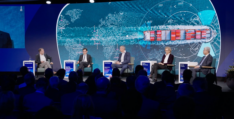Highlight video in an confrere - Five people on stage in suits converse during a panel. A digital map with a ship is displayed behind them. The setting is formal.
