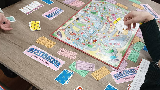 A lively board game club gathering with players of all ages enjoying various games. Some children are playing pick-up sticks, while older members engage in quiz challenges and a London-themed taxi board game called *Destination*. A table showcases an impressive collection of 150 board games, with Duo magnets as a new favorite. A welcoming atmosphere encourages donations of unwanted board games, ensuring every piece finds a new home. The club meets every Thursday at 6:00 PM, with plans for Saturday morning sessions.