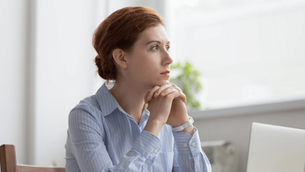 Focused executive woman reflecting at desk, hands clasped under chin, looking out window with thoughtful expression.
