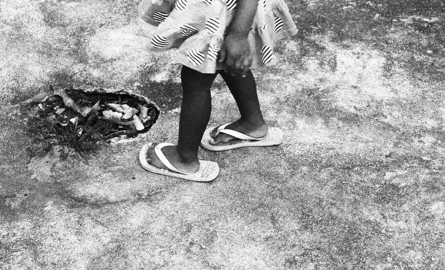 Créditos Manoel Peçanha. Menina diante de um buraco em meio ao chão acimentado. Ela está com vestido e uma Havaianas maior do que seu pé. No buraco, resíduos, matinhos, pedrinhas. Ao fundo, o chão acimentado cheio de manchas. Foto em P&B (preto e branco). Fim da AD.
