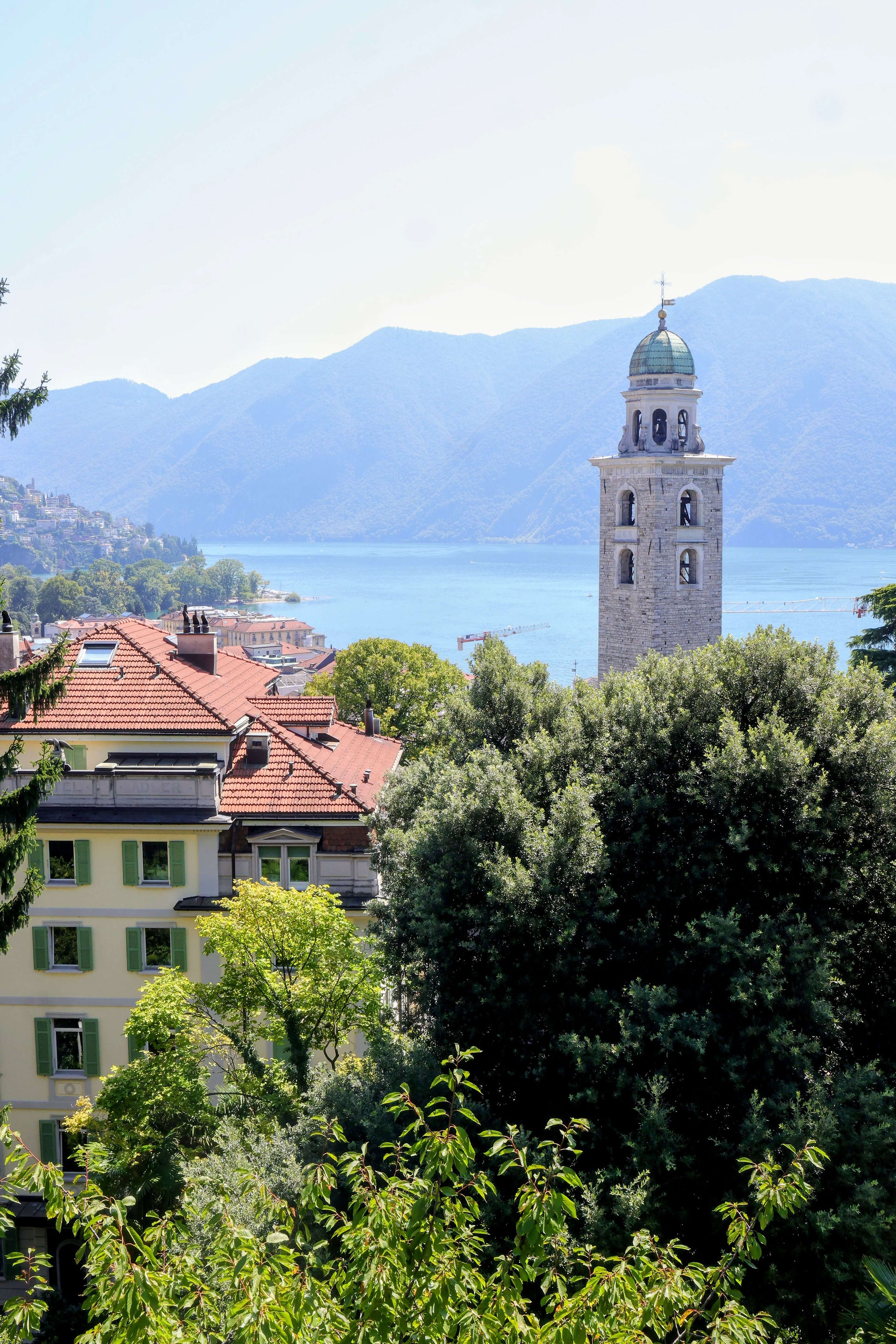 Blick über Dächer einer Stadt auf einen See und ein Bergpanorama mit einem Kirchturm im Vordergrund.