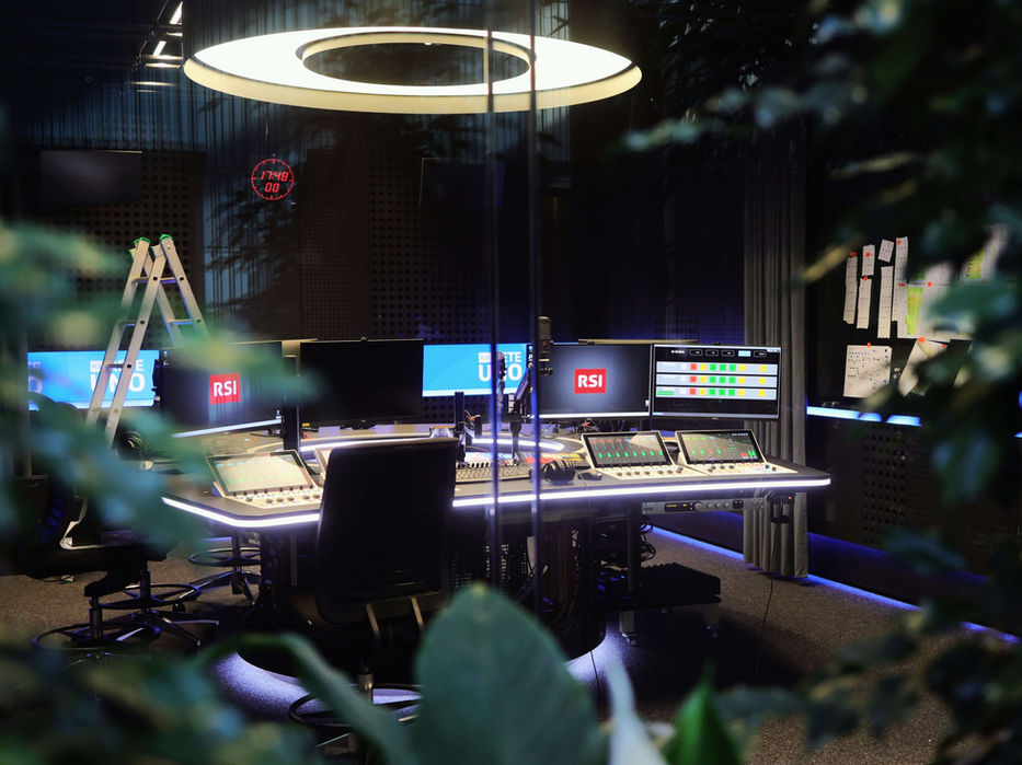 Regieraum mit großem Ringlicht an der Decke, Mischpult und mehreren Monitoren zur Steuerung und Überwachung von Audio‑ und Produktionssignalen.