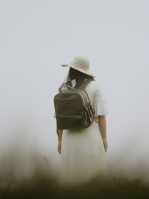 Woman wearing a hat and backpack hiking through a foggy outdoor field.