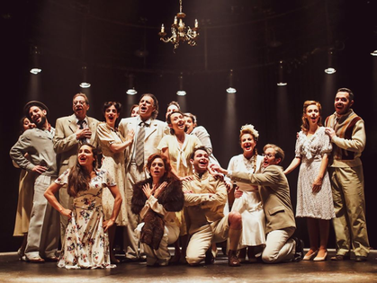 A group of people in vintage attire sing under spotlight in a theater setting. Warm lighting and chandelier create a nostalgic mood.