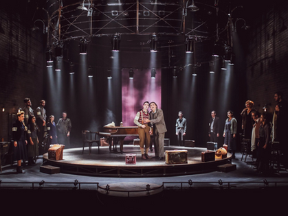 A group of actors in vintage attire stand on a stage with suitcases, under dramatic lighting, evoking a tense, theatrical atmosphere.