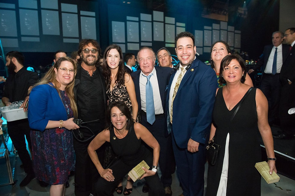 A group of people smiling in formal attire at an event with a blue-lit background. A waiter carries drinks on the left. Mood is cheerful.