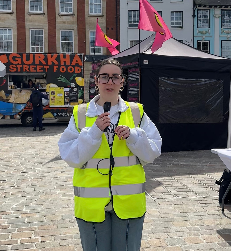 A person in a yellow vest holds a microphone in front of food stalls with vibrant signs and pink flags in a street market setting.