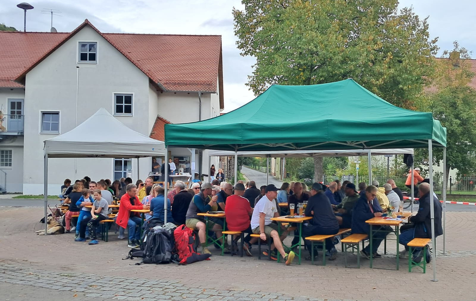 Viele Menschen sitzen unter grünen und weißen Pavillons an Biertischen vor einem Haus; links liegt ein Hund, gesellige Stimmung.