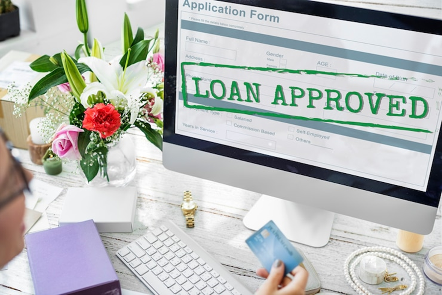 Person holding a credit card views a computer screen with "LOAN APPROVED." Desk has flowers, keyboard, purple notebook, and watch.