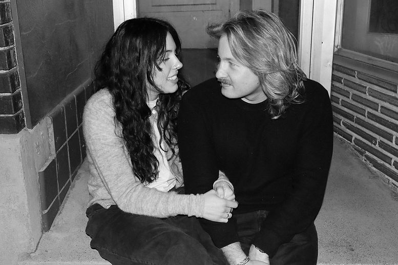 A photograph of a couple sitting on the front steps of an abandoned building, looking at each other. 