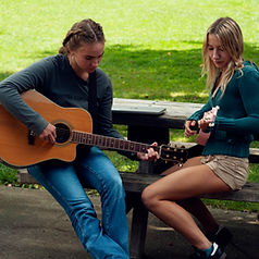Two female high school students at Summerfield Waldorf School are sitting at a picnic table playing a song together. The girl on the left is playing a guitar and the girl on the right is playing a pink ukulele.