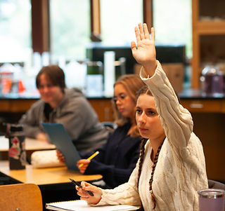 A female high school student at Summerfield Waldorf School is raising her hand to ask a question in class. She is wearing a white sweater and has long braids. Two classmates look on in the background.