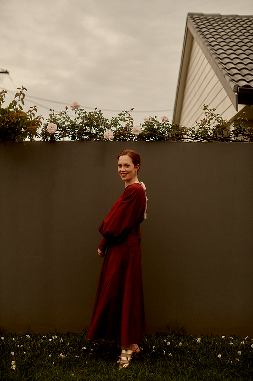Georgia Melville, soprano, smiling over her shoulder in a crimson long-sleeved dress. Photo by Saskia Wilson. Taken on Gadigal land in Sydney, Australia.