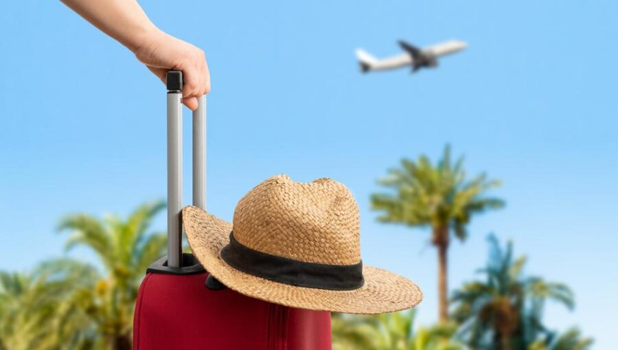 Close-up of a red travel suitcase and a straw hat with a plane flying overhead in a sunny sky.