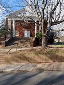 The front of a renovated red brick church buidling with white columns, a white roof, and steps leading to the entrance, partially covered by a large tree.