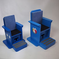 Two blue cardboard chairs with armrests stand side by side.  Each is mounted in a base that extends a few inches to the back and sides.  The taller chair has a beach ball painted on its side.  Each chair has a blue-covered cushion, black yoga matting on the seat, and a footrest with non-skid strips.