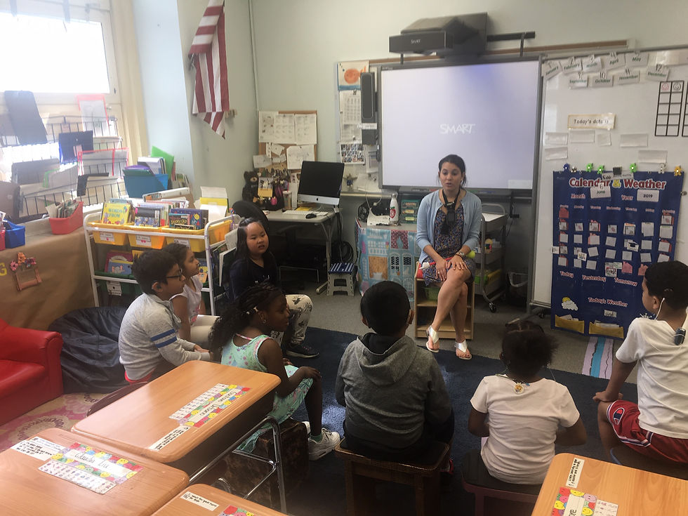 In her classroom, teacher Hayley Dick sits with her students in front of her in a semicircle. Behind her hangs a large projector screen and a white board with black writing on it, along with other classroom projects.