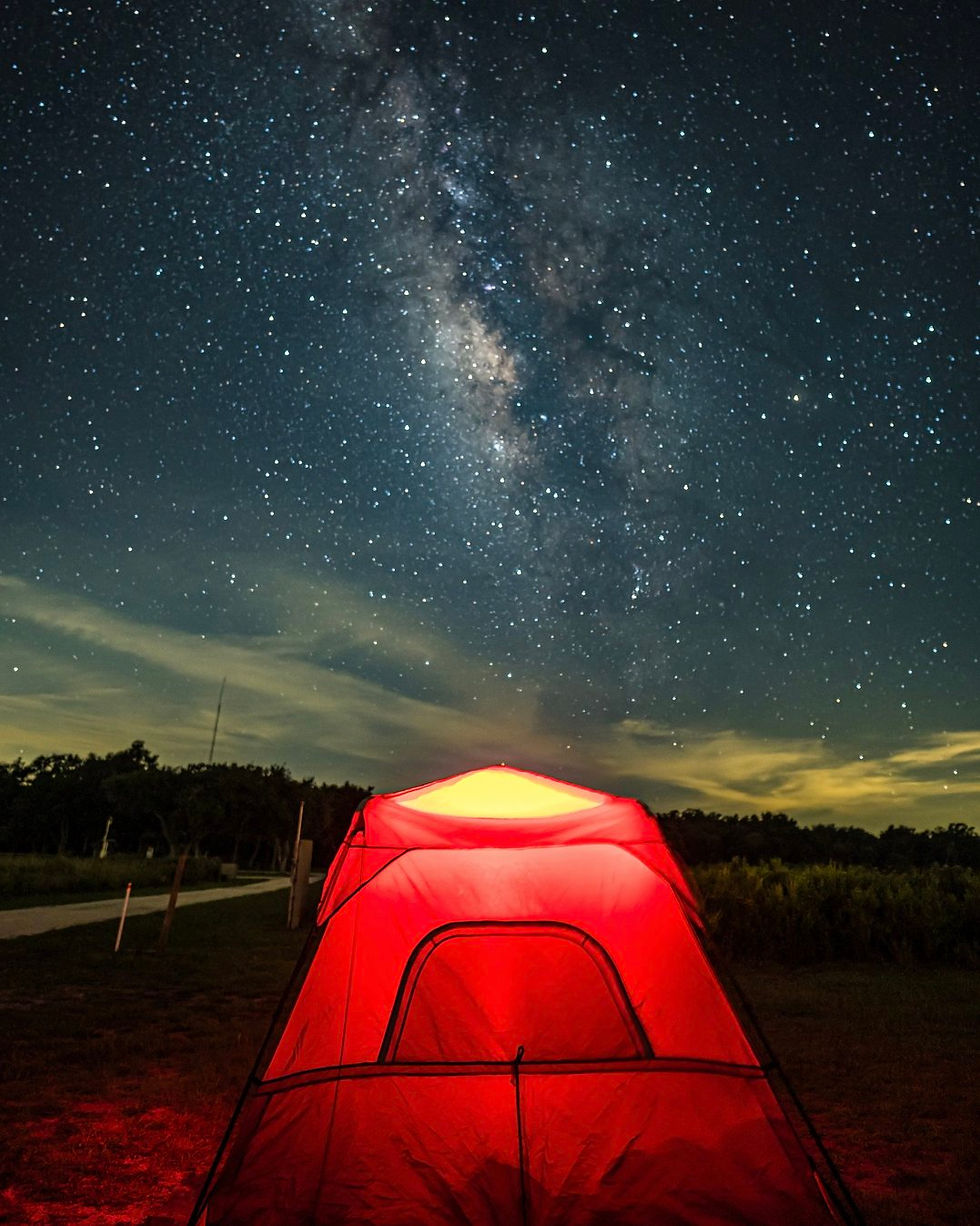 Glowing red tent in a dark field under a star-filled Milky Way night sky