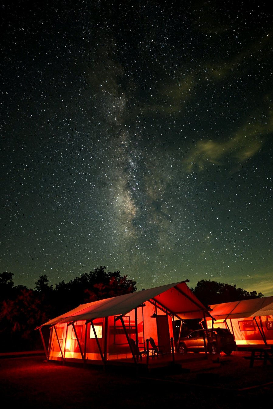 Milky Way over glowing red campsite tents under a star-filled night sky.