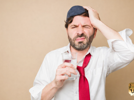 Man in a white dress shirt and loosened red tie pressing his hand to his head while holding a glass of water, experiencing hangover symptoms the morning after