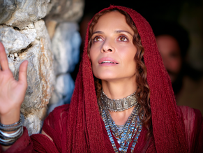 Woman in a red shawl touches a stone wall, looking up with a serene expression. She wears silver jewelry. Background is dimly lit.