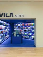 Livraria da Vila Artes，米白色店面从商场木地板旁切出深蓝入口，书架与中央展台在门框中形成完整的第一眼。
Livraria da Vila Artes, An off-white storefront cuts a deep-blue opening beside the mall's timber floor, framing shelves and the central display in a single first view.