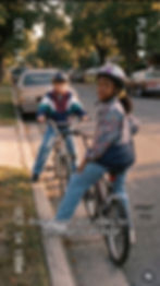 Two children on their bicycles pause at a street curb and look toward the camera. They wear bicycle helmets. In the background are cars parked in the distance.