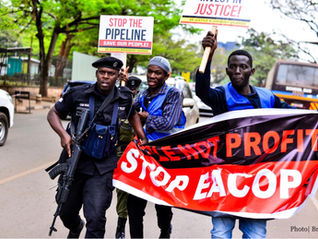 An armed official grabs a banner held by protesters whose signs say "Stop the Pipeline" and "Stop EACOP".