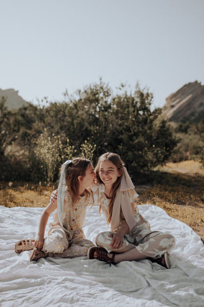 Two sisters sit together on a white blanket at Vasquez Rocks, one whispering a secret into the other’s ear, sharing a sweet sibling moment. Los Angeles family photography specializing in natural interactions.