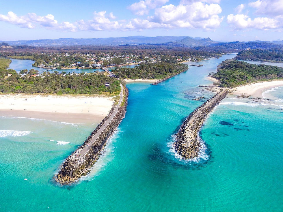 Aerial view of the vibrant Brunswick Heads 
turquoise coastline with breakwaters, sandy beaches, lush greenery, and a distant town under a blue sky with clouds.
