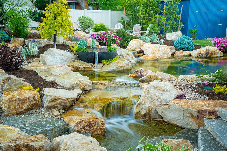 Backyard pond and waterfall landscaping with rock features, flowing water, aquatic plants, and a fountain bowl surrounded by flowers
