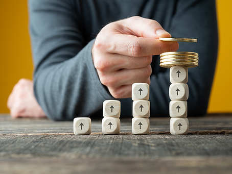 Hand stacking coins on blocks with upward arrows, wooden table, yellow background, gray sleeve visible, symbolizing financial growth.