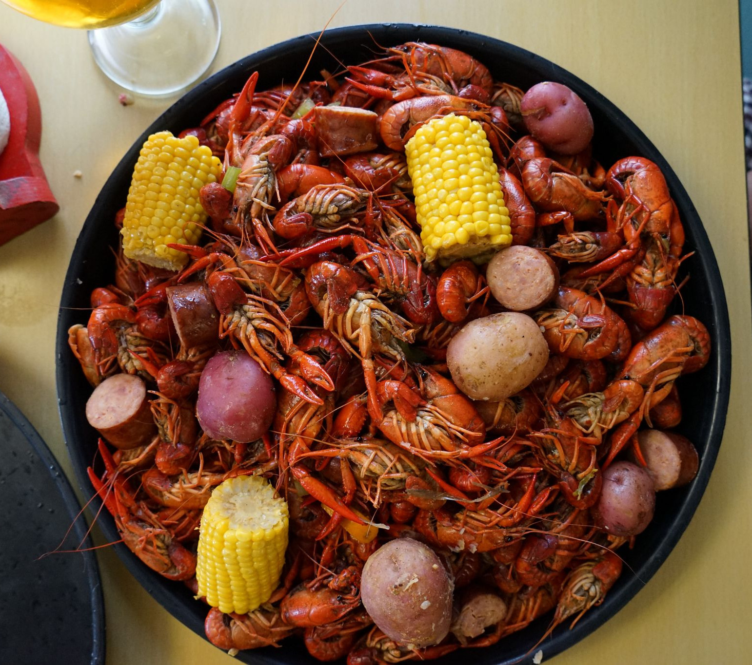 Top-down view of a black bowl piled with red crawfish, corn on the cob, potatoes, and sausage on a table, with a drink nearby