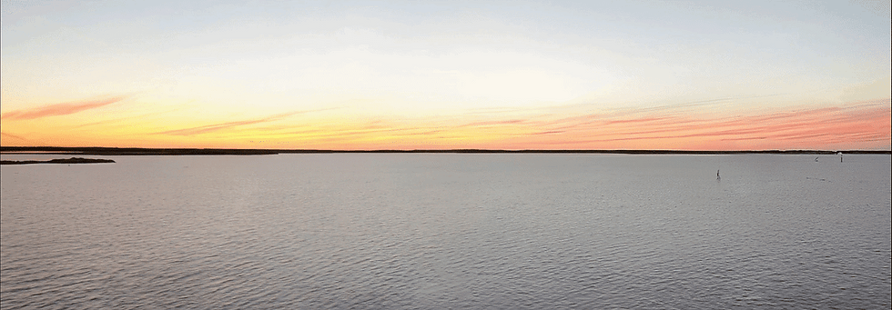 Calm wide lake at sunset, with pastel orange-pink sky and distant dark shoreline over rippling water.