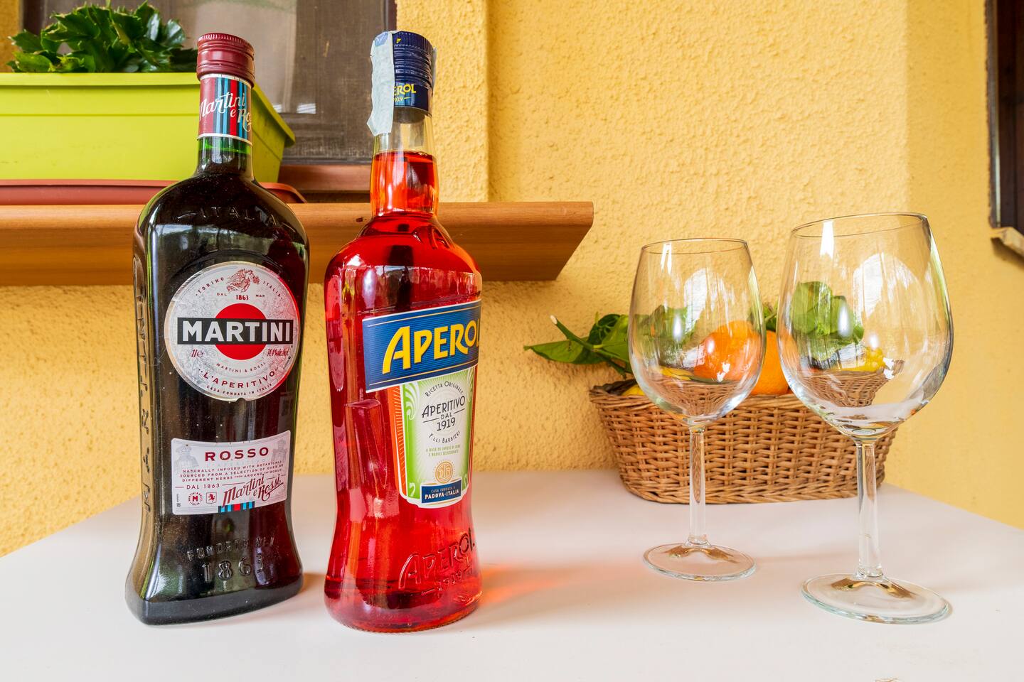 Bottiglie Martini Rosso, Aperol con bicchieri da vino su tavolo bianco.