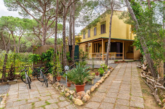 Edificio giallo con due piani tra pini, biciclette parcheggiate e sentiero giardino.