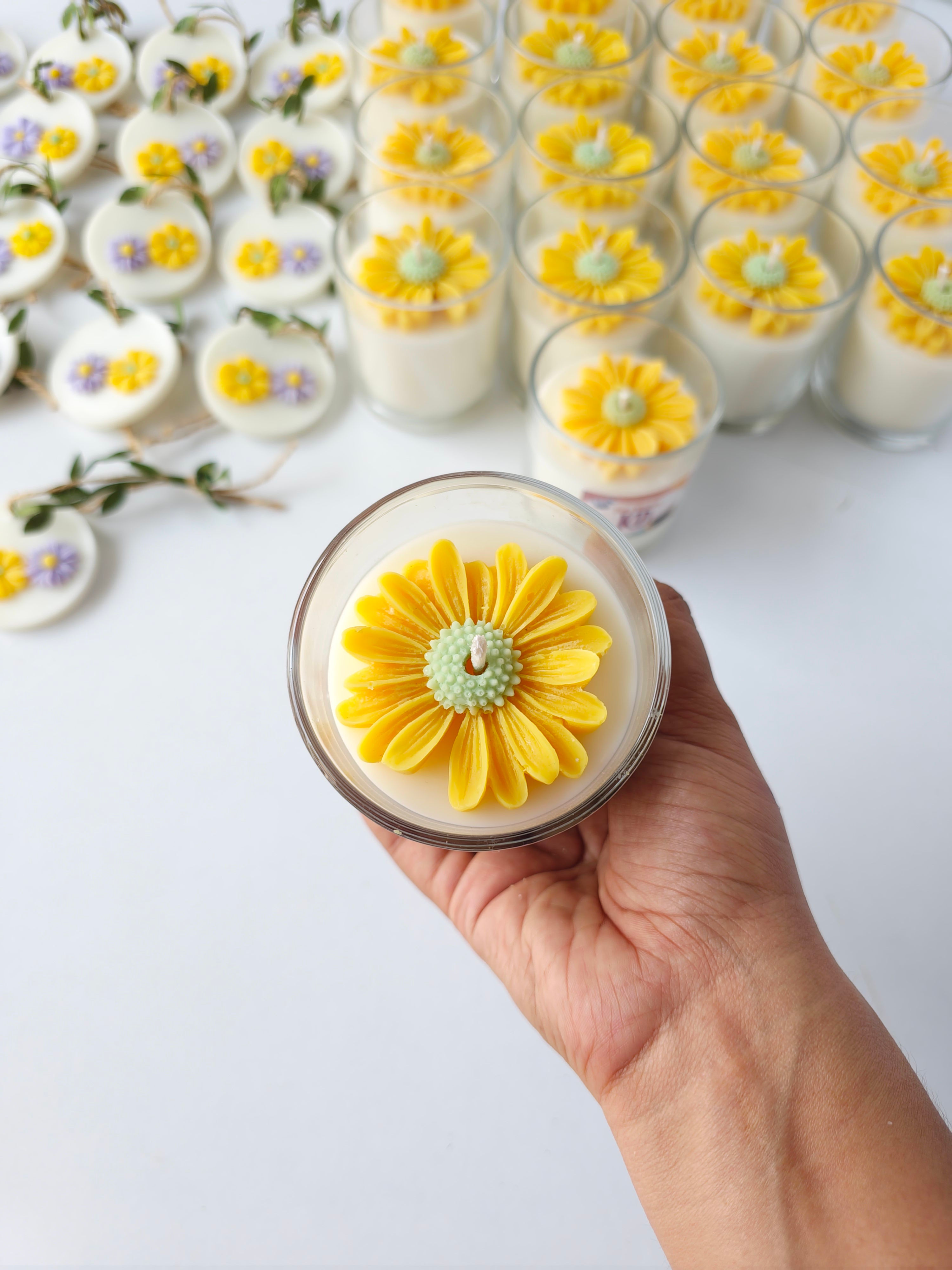 Hand holding a daisy flower topped scented soy wax candle, with rows of matching candles behind.