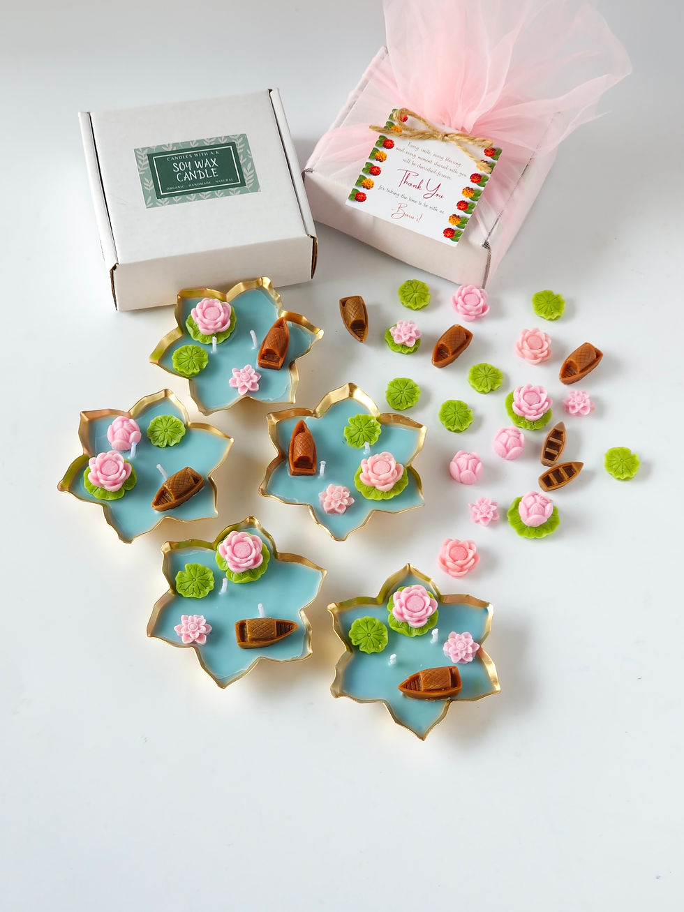 Scented soy wax lotus pond urli candles with pink lotus and boat toppers, beside gift boxes.