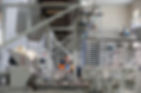 Man in an orange safety vest stands in a modern bread factory among machines with dough. Industrial setting with metal structures, bright lighting.