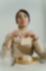 Person wearing a beige sweater holds a slice of bread solemnly. A plate with UPF white sandwich squares is on a white table, set with cutlery.