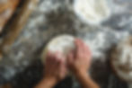 Hands kneading dough on a floured wooden table. Nearby are a bowl of flour, rolling pin, and baked bread. Rustic and textured setting. Making real bread at home as a healthier option