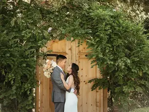 Bride and groom embrace in front of a wooden gate, surrounded by lush greenery. Bride holds a bouquet, creating a romantic mood.